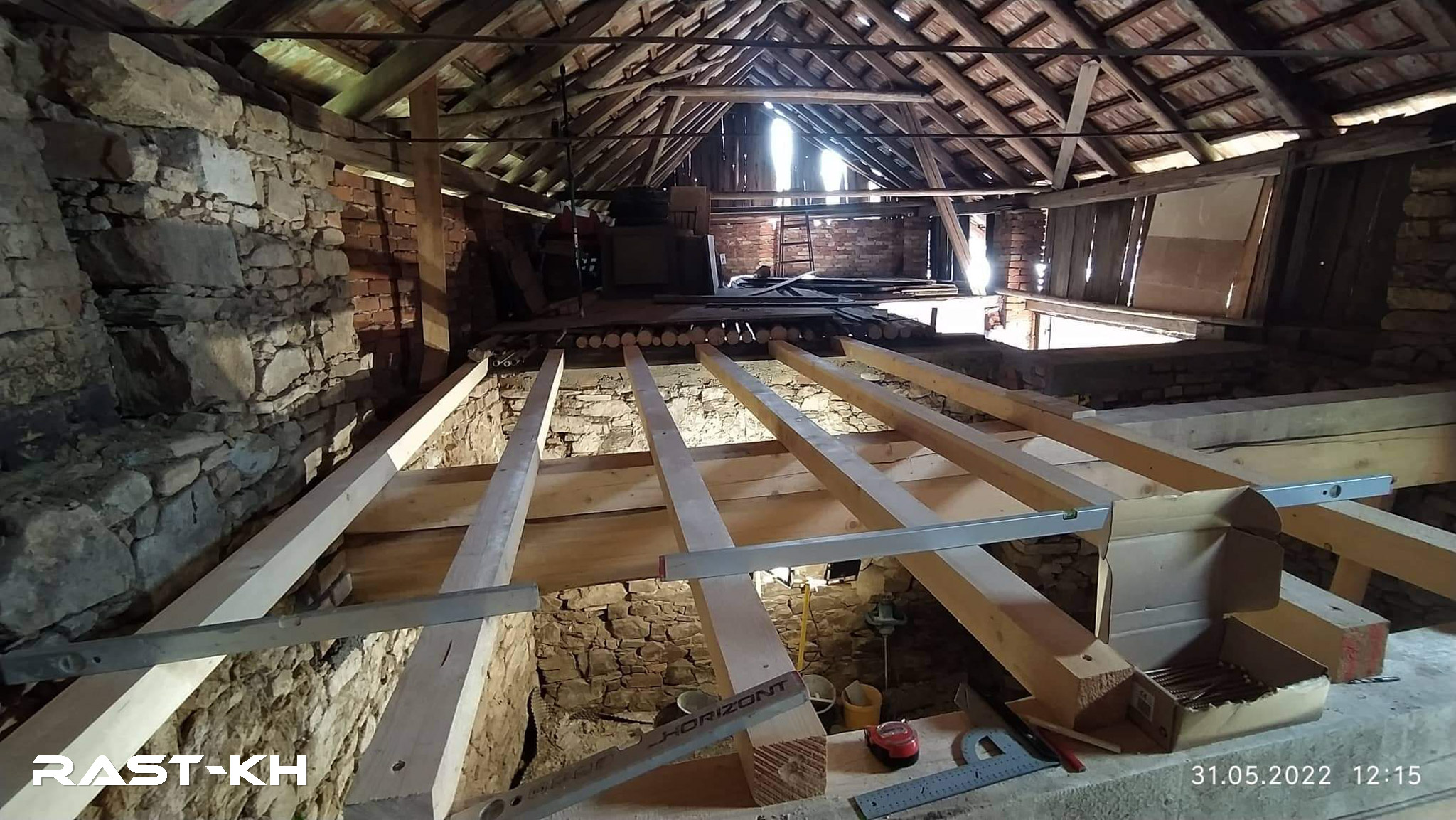 Rekonstrukce podkrovního prostoru, pohled na dřevěné trámy podlahové konstrukce ve starém kamenném domě. Trámy jsou nově instalované mezi kamenné stěny, nad nimiž se rozprostírá původní dřevěná střešní konstrukce s odkrytými cihlami a trámovím.